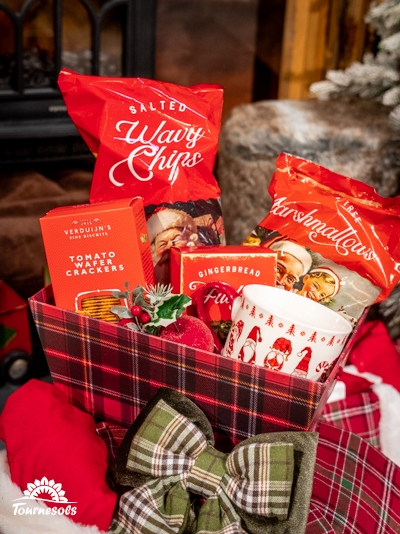 Panier garni avec chips, crackers, guimauves, et tasse de Noël sur fond festif.